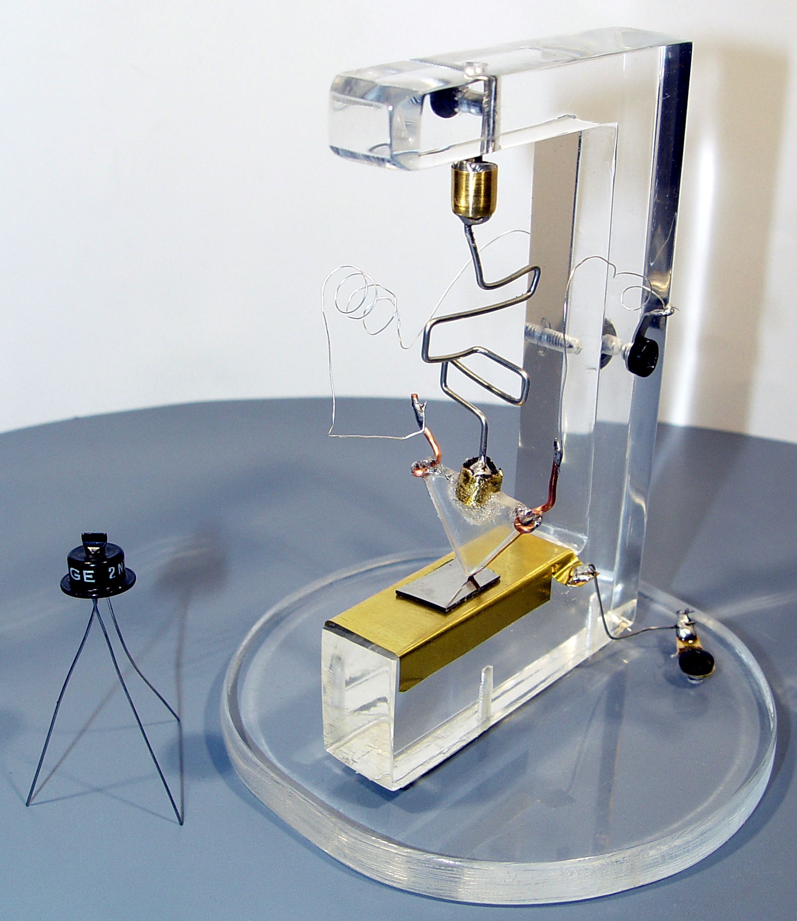 A replica of the first point-contact transistor beside a small 2N43 transistor on a pale background.